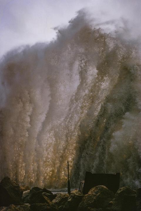 A large wave crashes against the Scarborough coast, with rocks and a pole in the foreground.