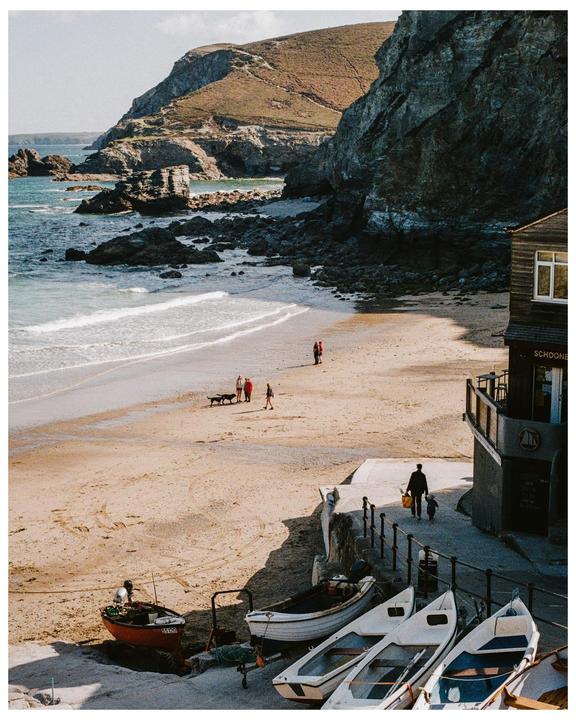 A sandy beach with people and boats is nestled between rocky cliffs and a building.