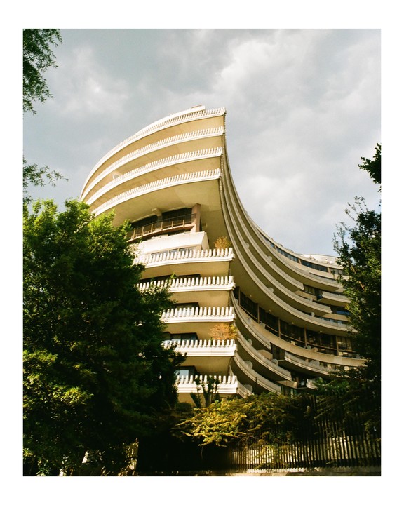 A curved building with balconies is partially obscured by trees under a cloudy sky.