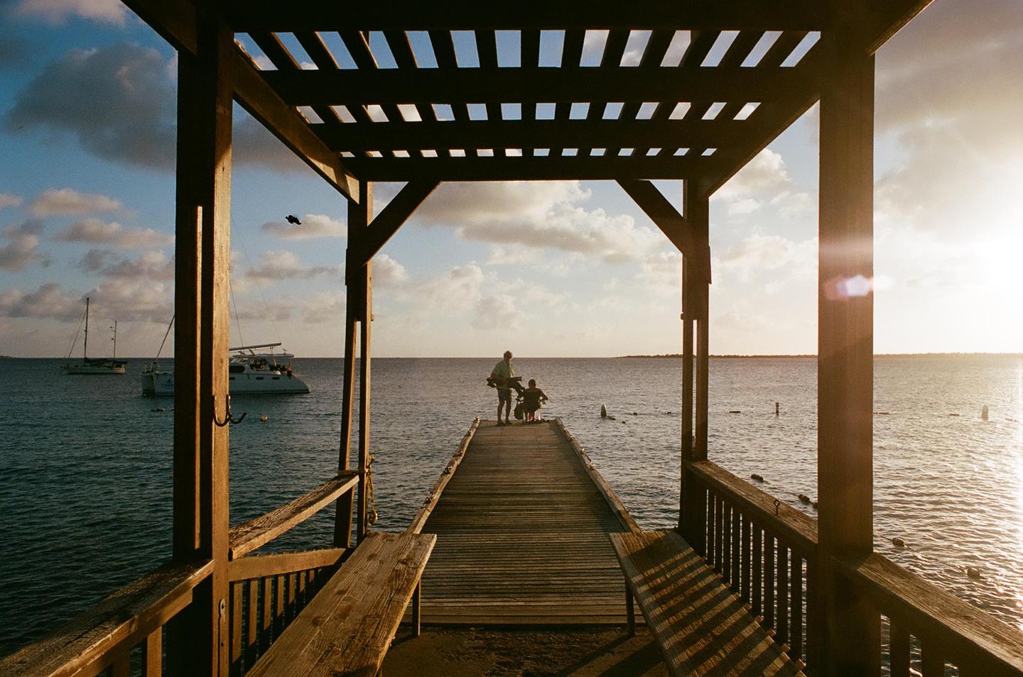 A man and a child stand on a wooden pier looking out at the ocean with boats in the distance under a cloudy sky at sunset.