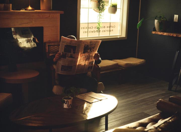 A person sits in a chair reading a newspaper in a dimly lit room with sunlight streaming through a window.