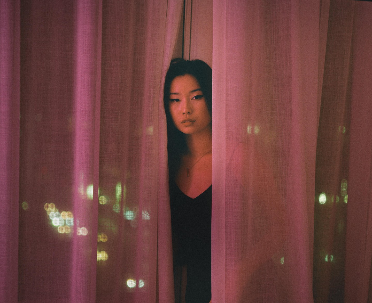 A woman peeks through pink curtains with city lights visible in the background.