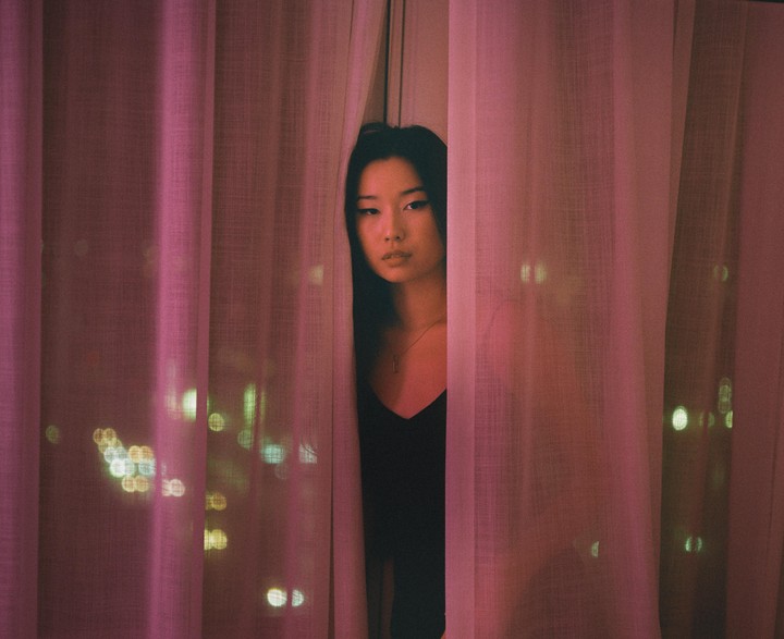 A woman peeks through pink curtains with city lights visible in the background.