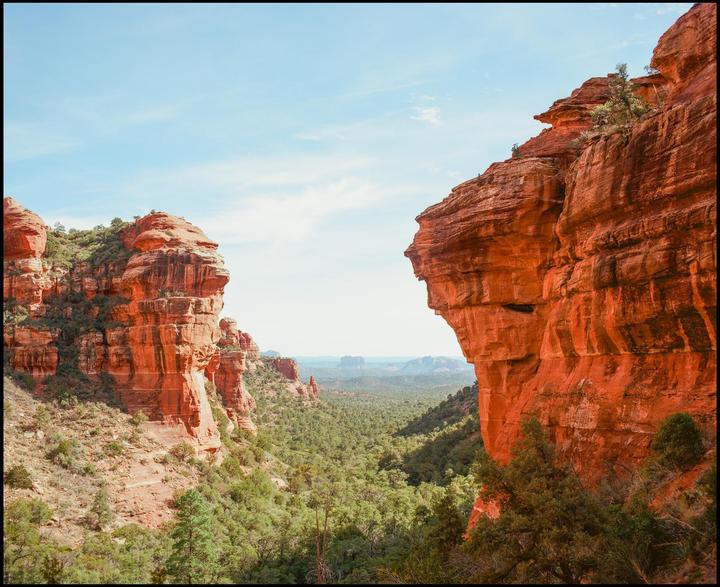 A wide view shows a valley filled with green trees and bushes, flanked by two large red rock formations under a bright sky.