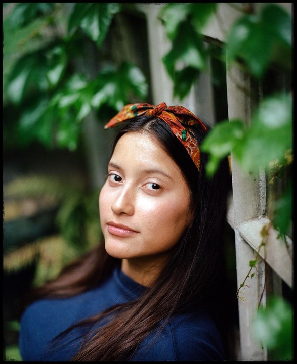 A woman with a patterned headband and a blue shirt looks at the viewer, framed by green foliage.