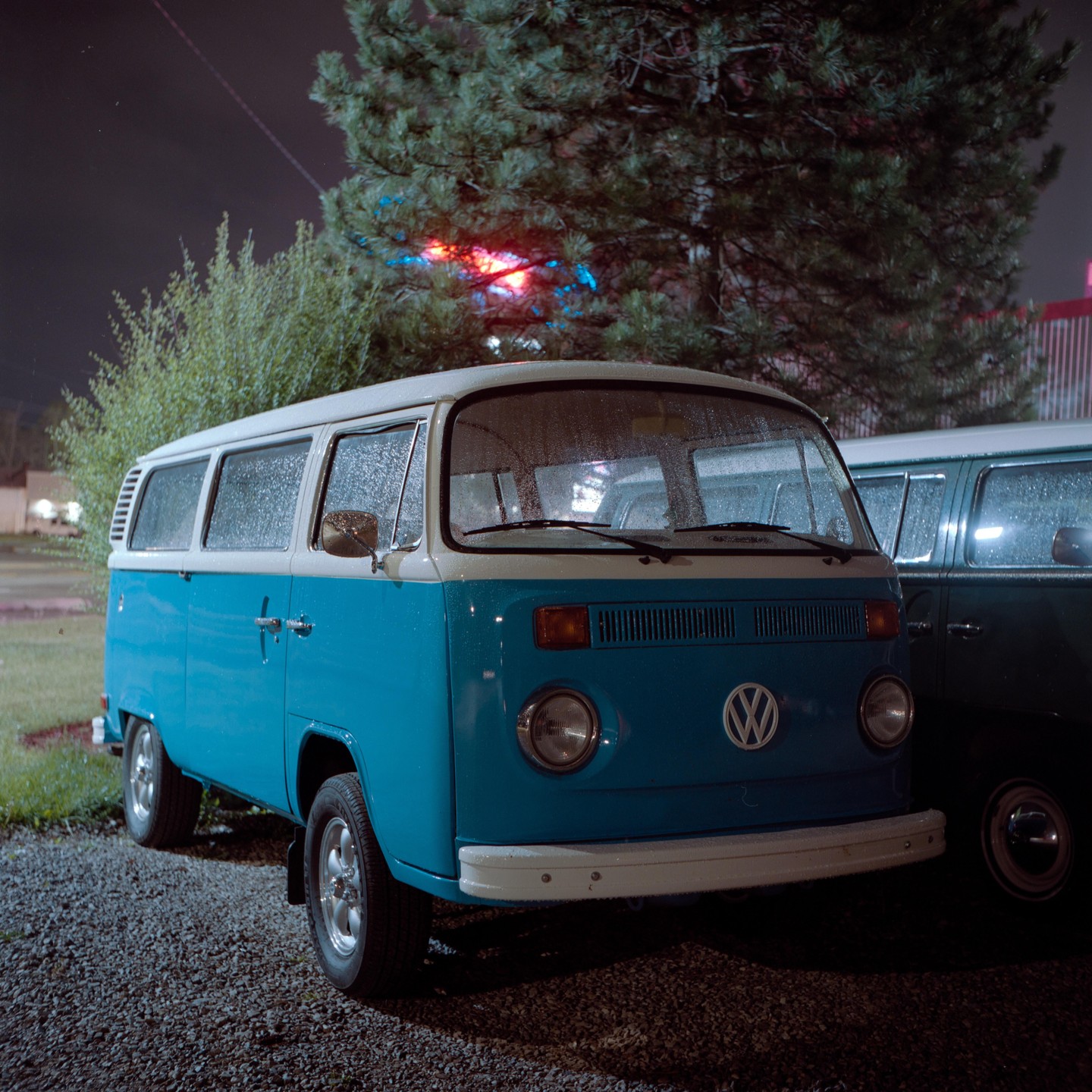 A blue and white van is parked on a gravel lot at night, with another van visible behind it.