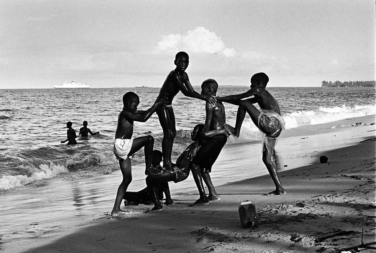 A group of boys play on a beach with a large ship visible in the distance.