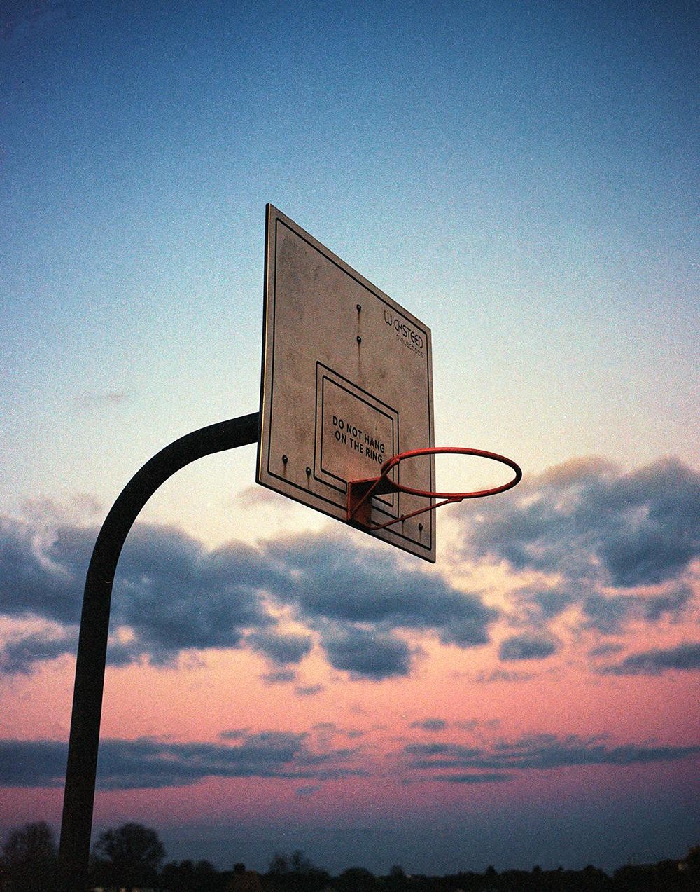 A basketball hoop and backboard are silhouetted against a colorful sunset sky with clouds.