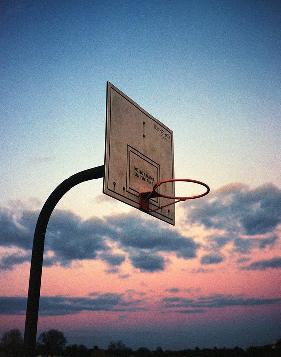 A basketball hoop and backboard are silhouetted against a colorful sunset sky with clouds.