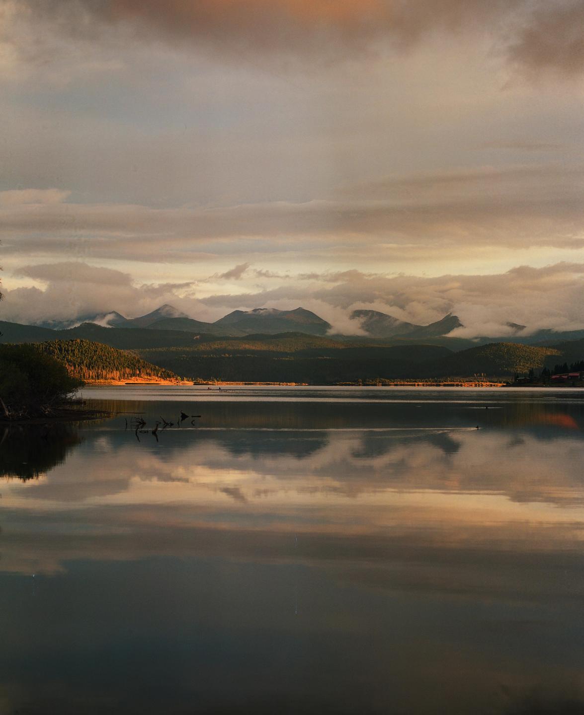 A lake reflects the cloudy sky and mountains with trees on the shore.