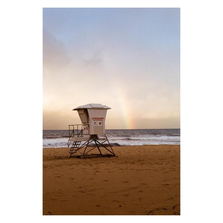 A lifeguard stand sits on a sandy beach with a rainbow visible over the ocean.