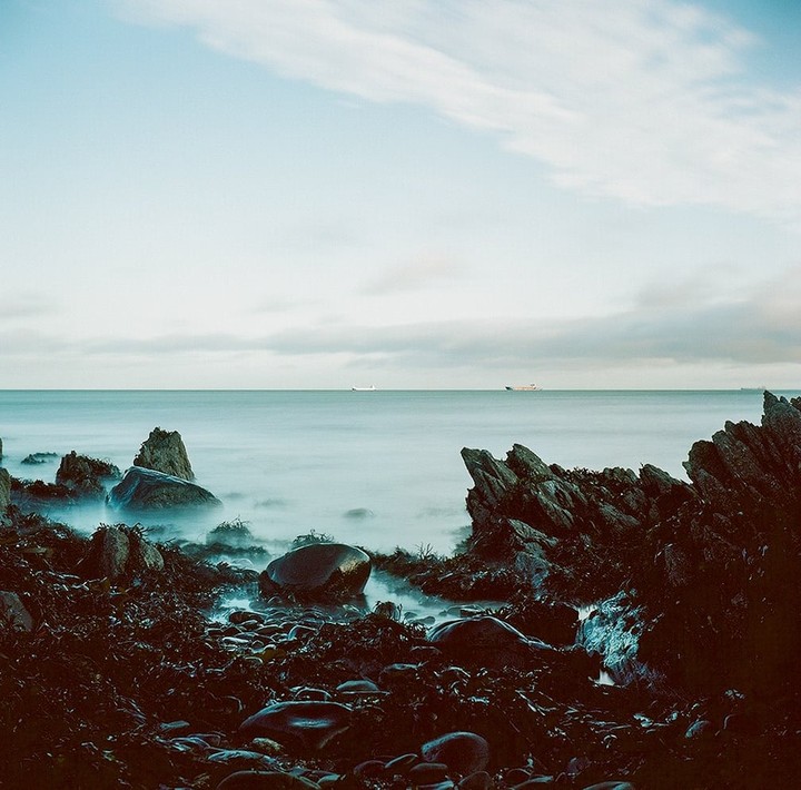 A long exposure shot captures the smooth surface of the sea with rocks and seaweed in the foreground and two ships on the horizon.
