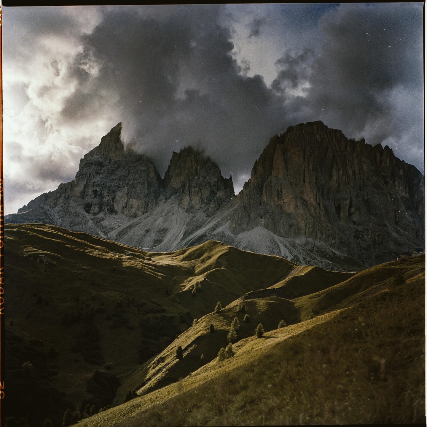 A dramatic landscape shows mountains under a cloudy sky with sunlit rolling hills in the foreground.