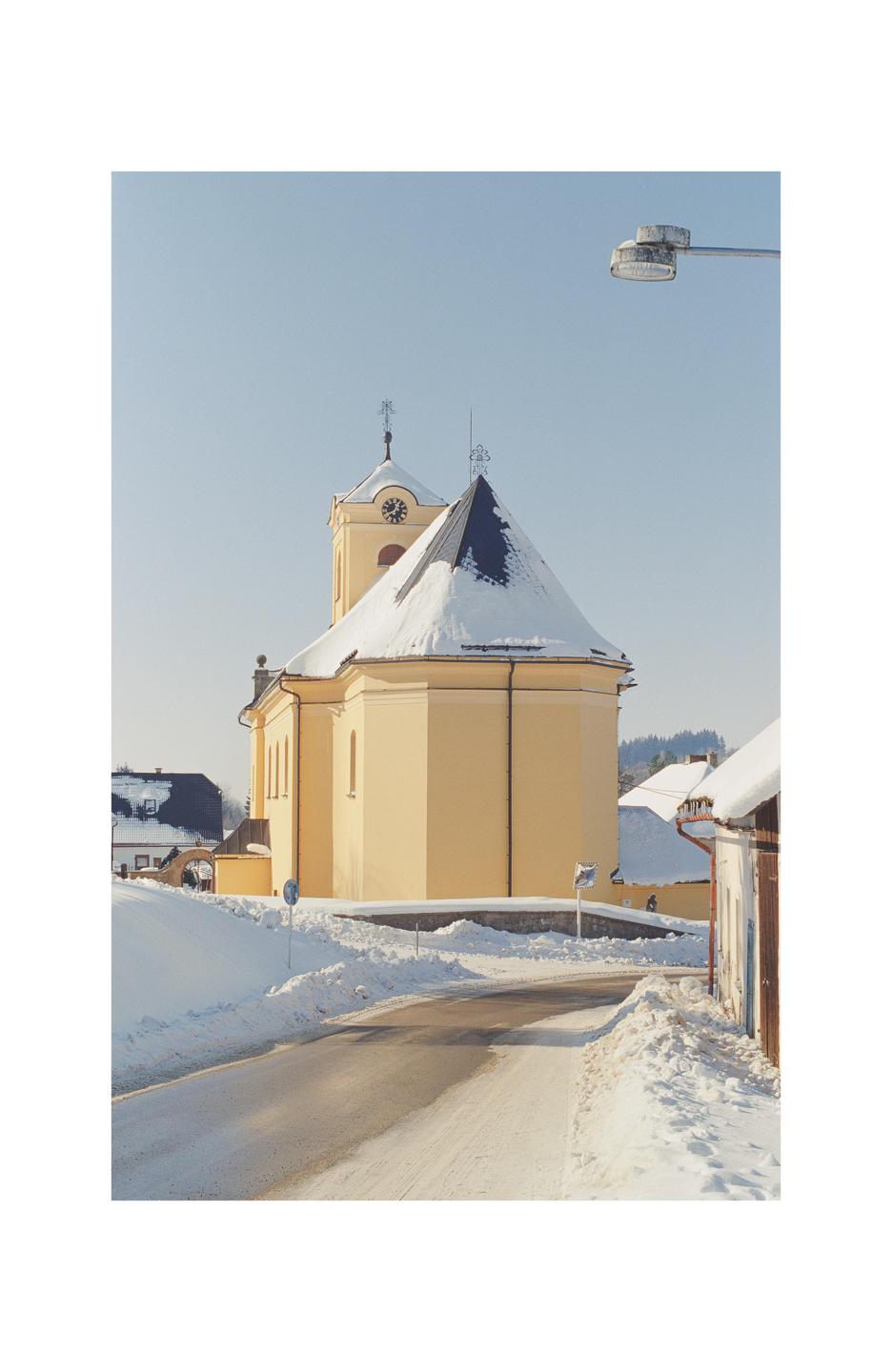 A yellow church with a snow-covered roof stands beside a snowy road under a clear sky.