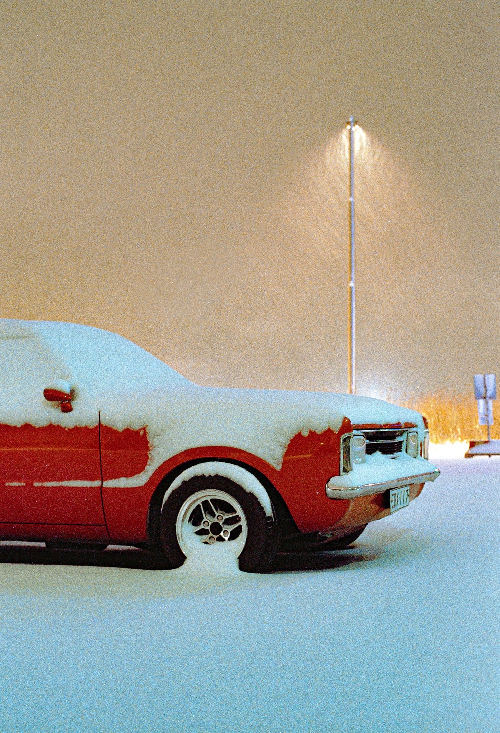A red car is covered in snow next to a street light at night.