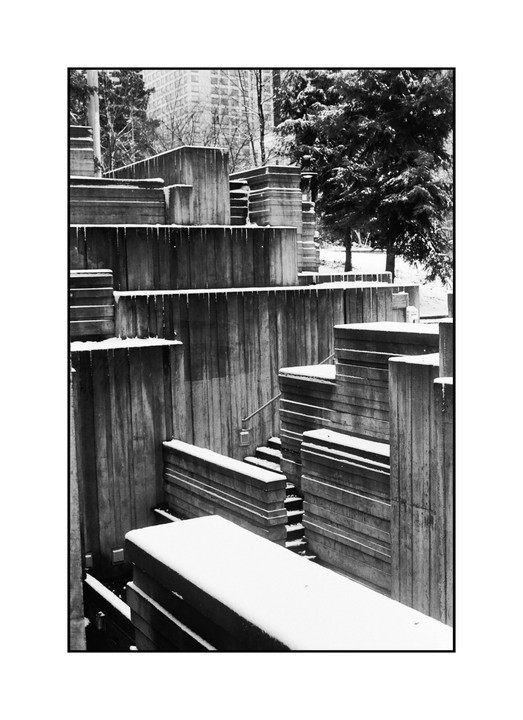 A concrete brutalist park with snow on its surfaces and icicles hanging from ledges is shown in black and white.