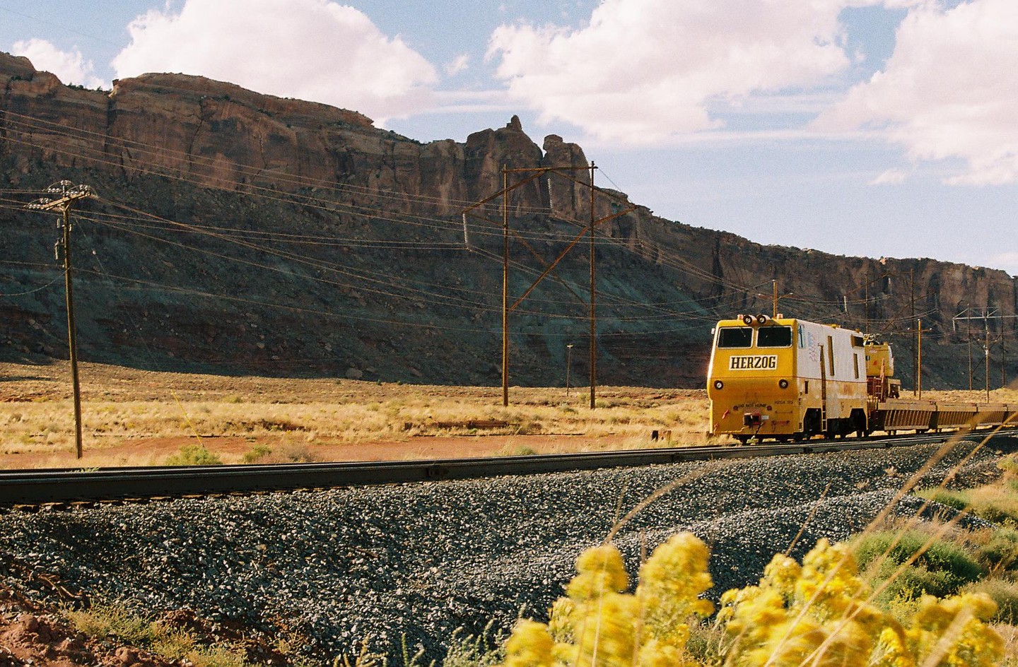 A yellow Herzog train travels along a railway track next to a large rock formation under a partly cloudy sky.