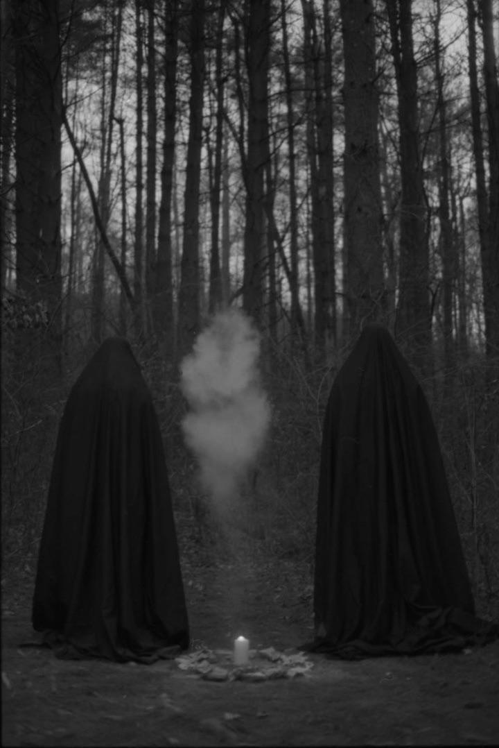 Two figures in dark cloaks stand in a forest with a lit candle and smoke between them.
