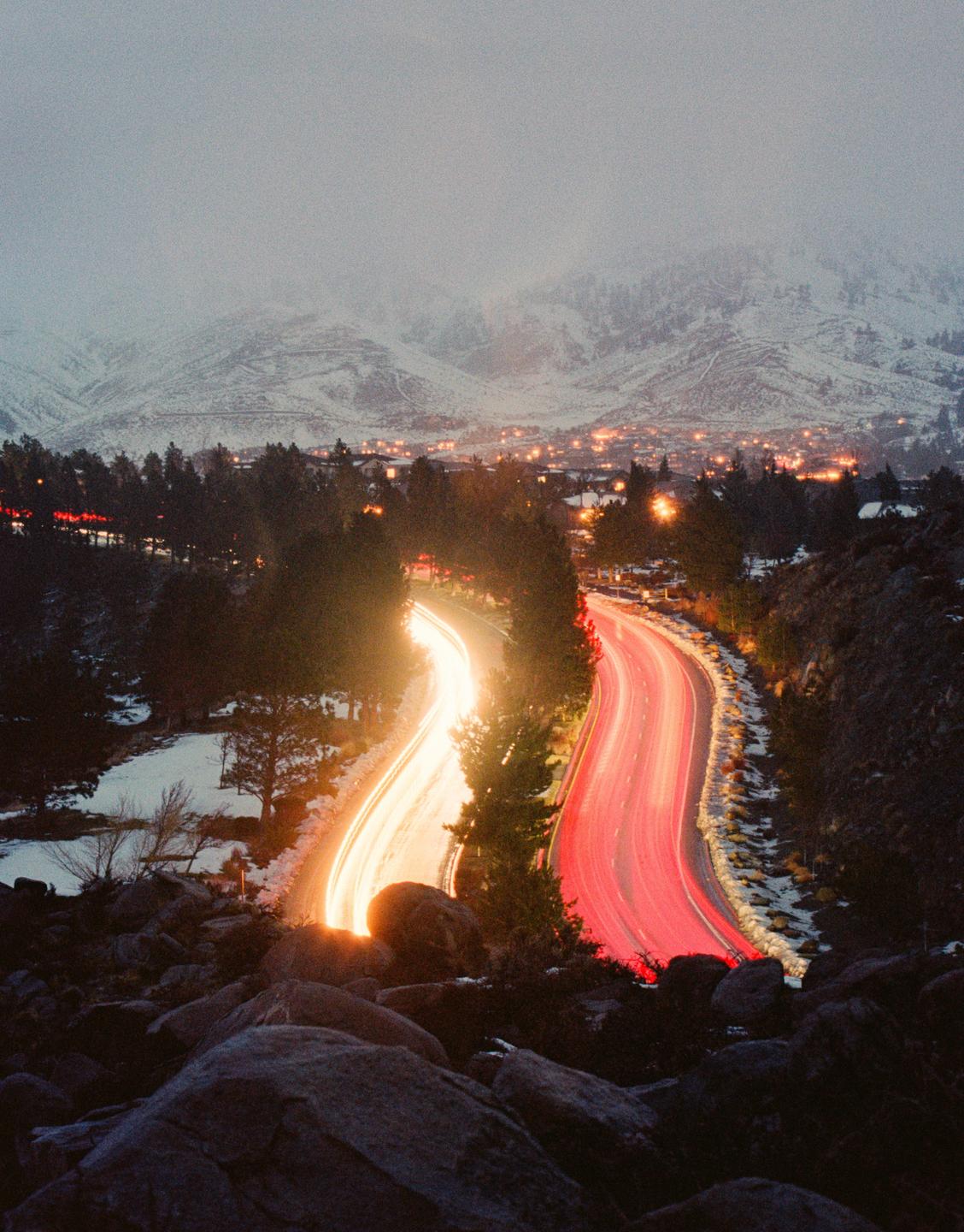 Light trails from vehicles on a winding road lead to a town nestled in a snowy mountain valley at night.