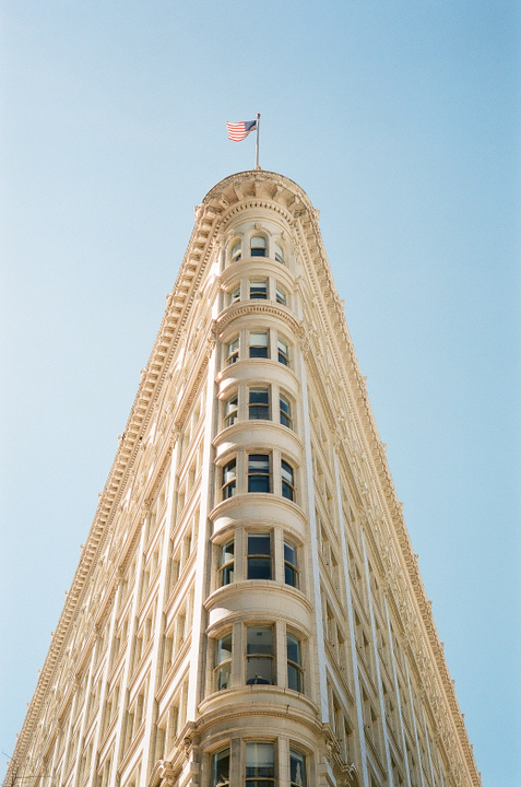 A low-angle view shows the ornate facade of the Phelan Building with an American flag flying from its rounded corner against a clear sky.