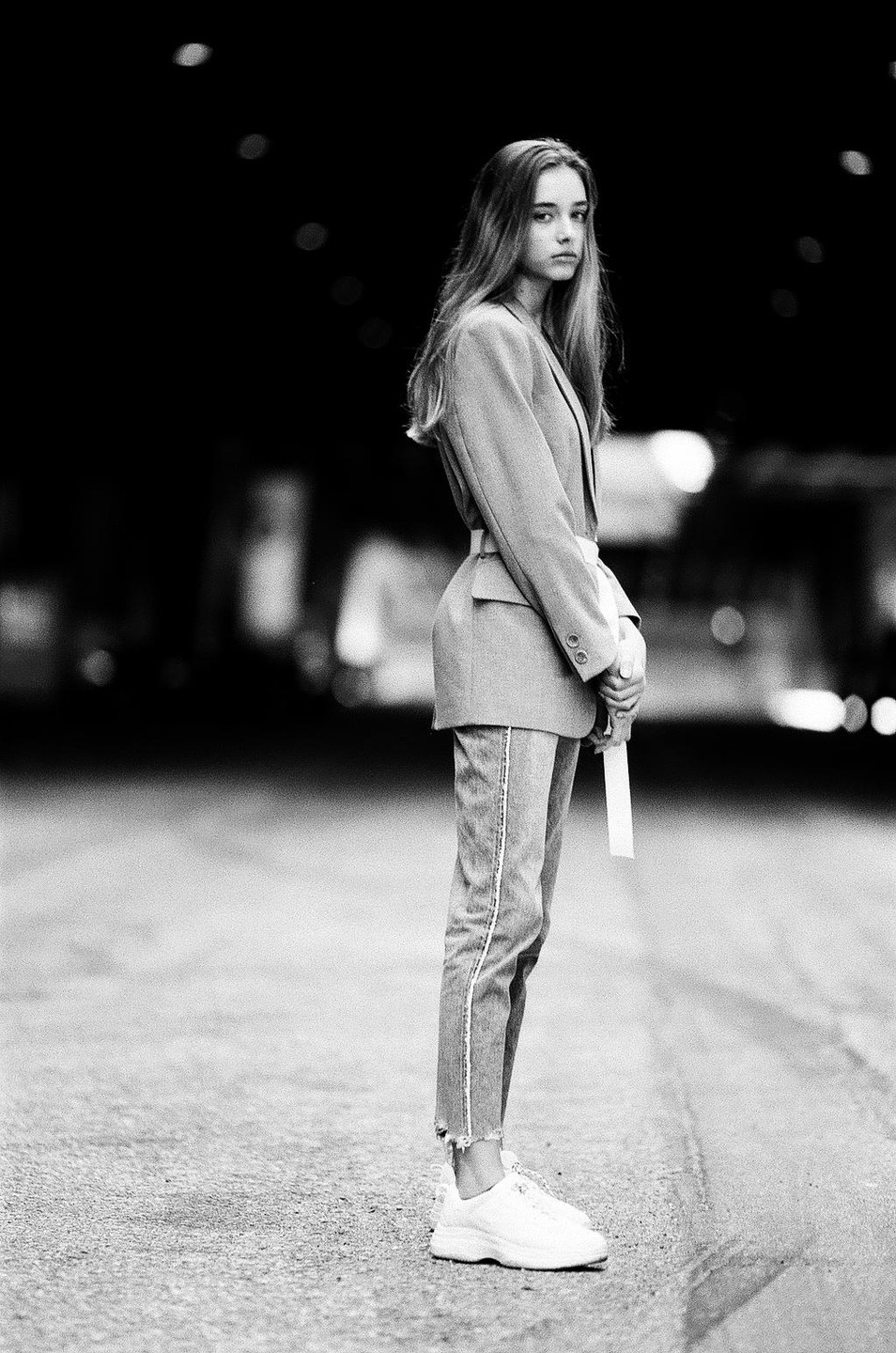 A young woman stands in the middle of a street at night, looking over her shoulder.
