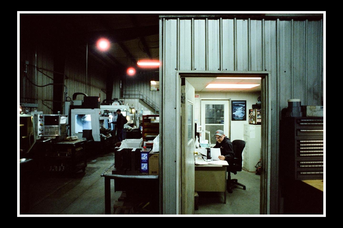 A man sits at a desk in an office next to a large industrial workshop with machinery and another man.