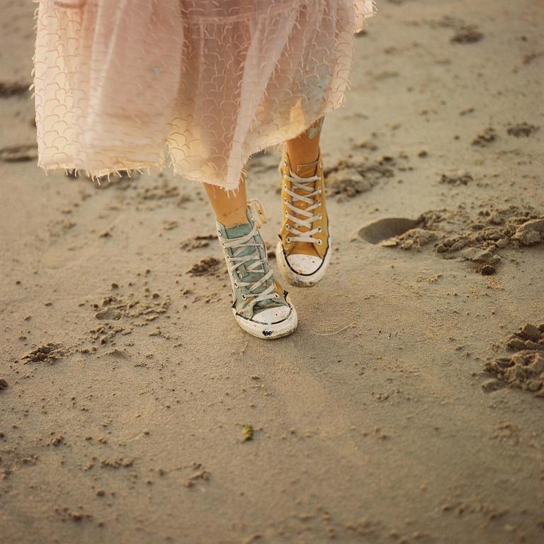 A person wearing a dress and two different colored sneakers walks on a sandy beach.