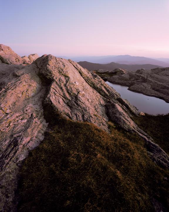 A small lake is nestled among rocky mountains with sparse vegetation under a soft pink and purple sky.