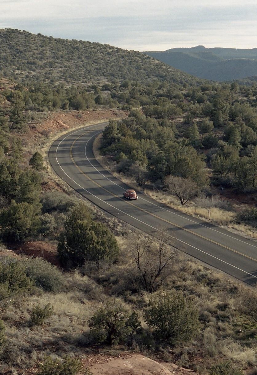 A red car drives along a winding road through a hilly, forested landscape under a cloudy sky.