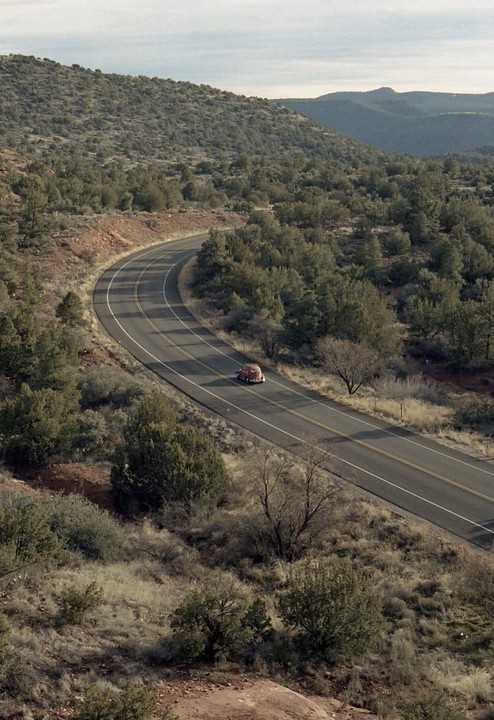 A red car drives along a winding road through a hilly, forested landscape under a cloudy sky.