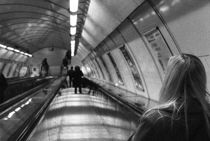 A woman with long hair stands on an escalator in a subway station, looking forward as other people ascend in the distance.