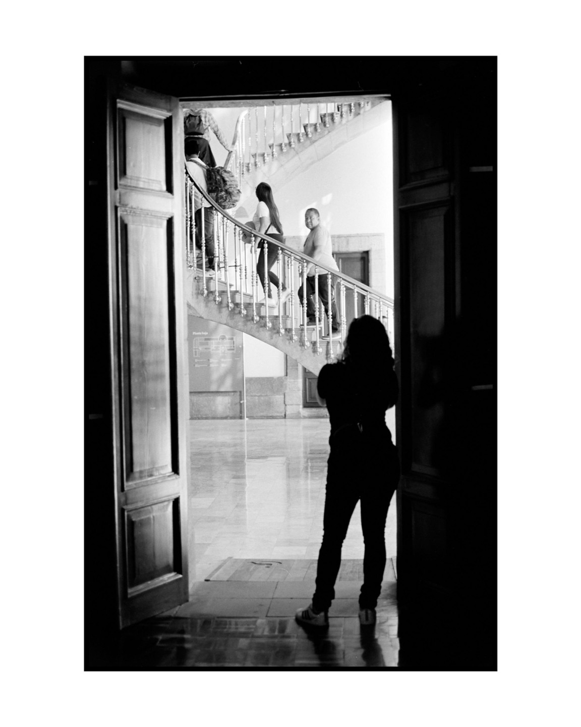 A person stands in a doorway, silhouetted against a bright interior with people on a curved staircase.
