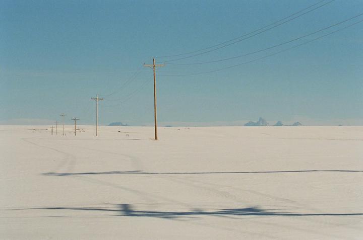 A vast, snow-covered plain stretches towards distant mountains under a clear sky, with a line of utility poles and their shadows extending into the distance.