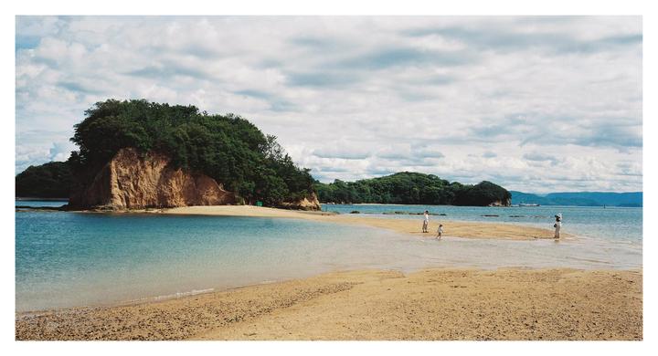 A narrow sandbar connects two islands with people walking on it, surrounded by blue water and green hills under a cloudy sky.