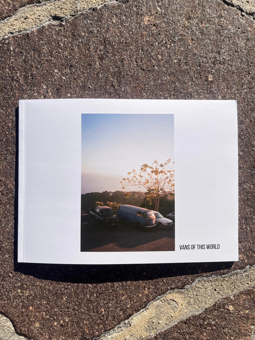 A white zine with a photograph of vans and a tree at sunset is resting on a stone surface.