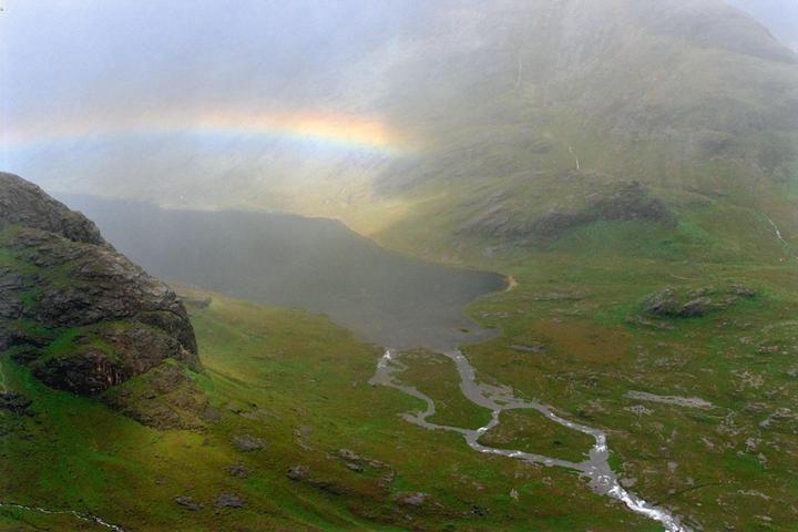 A rainbow arches over a lake and a valley with a stream flowing through it.