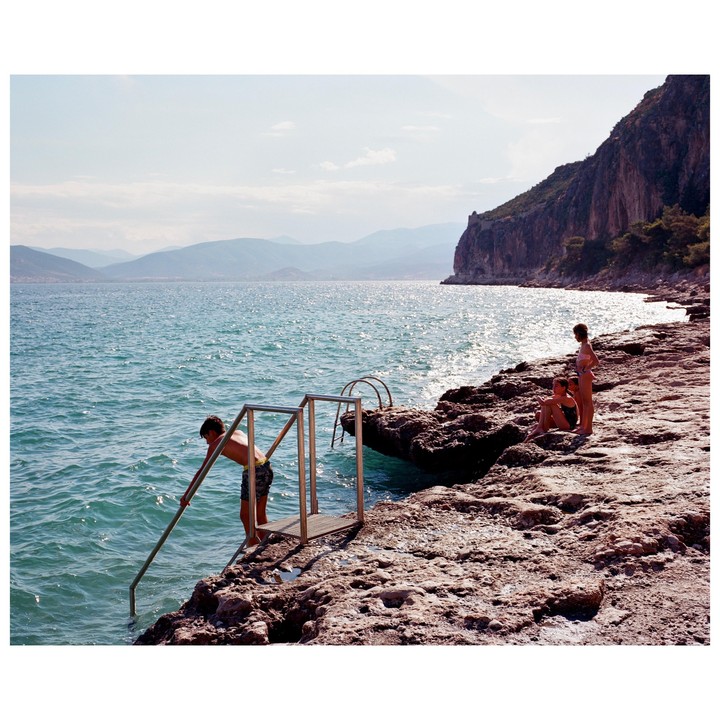 A boy descends a ladder into the sea from a rocky shore, while two children stand nearby.