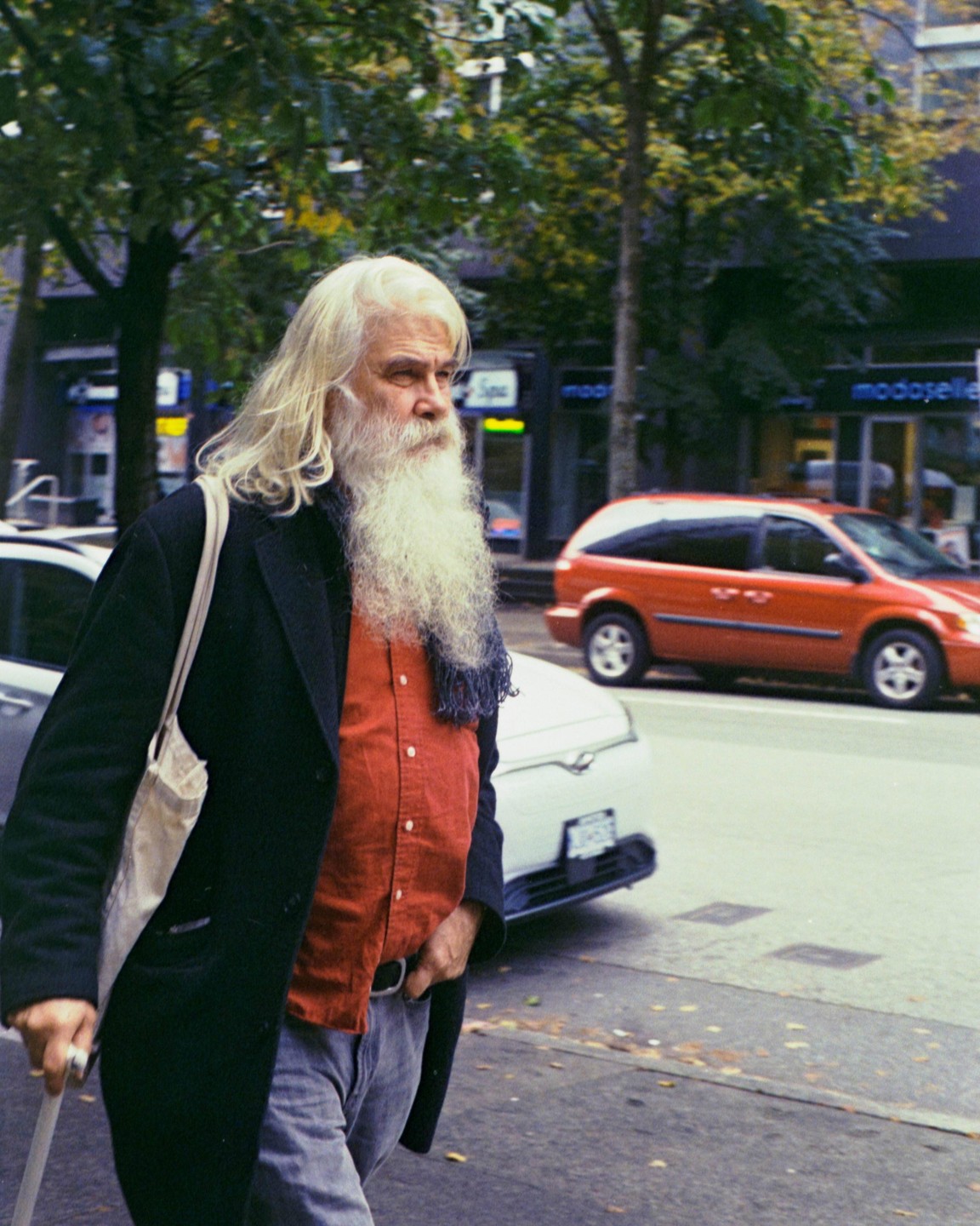 A man with a long white beard and long white hair walks on a city street.