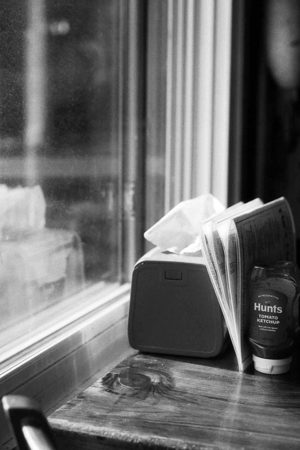 A tissue box, a menu, and a ketchup bottle sit on a wooden table next to a window.