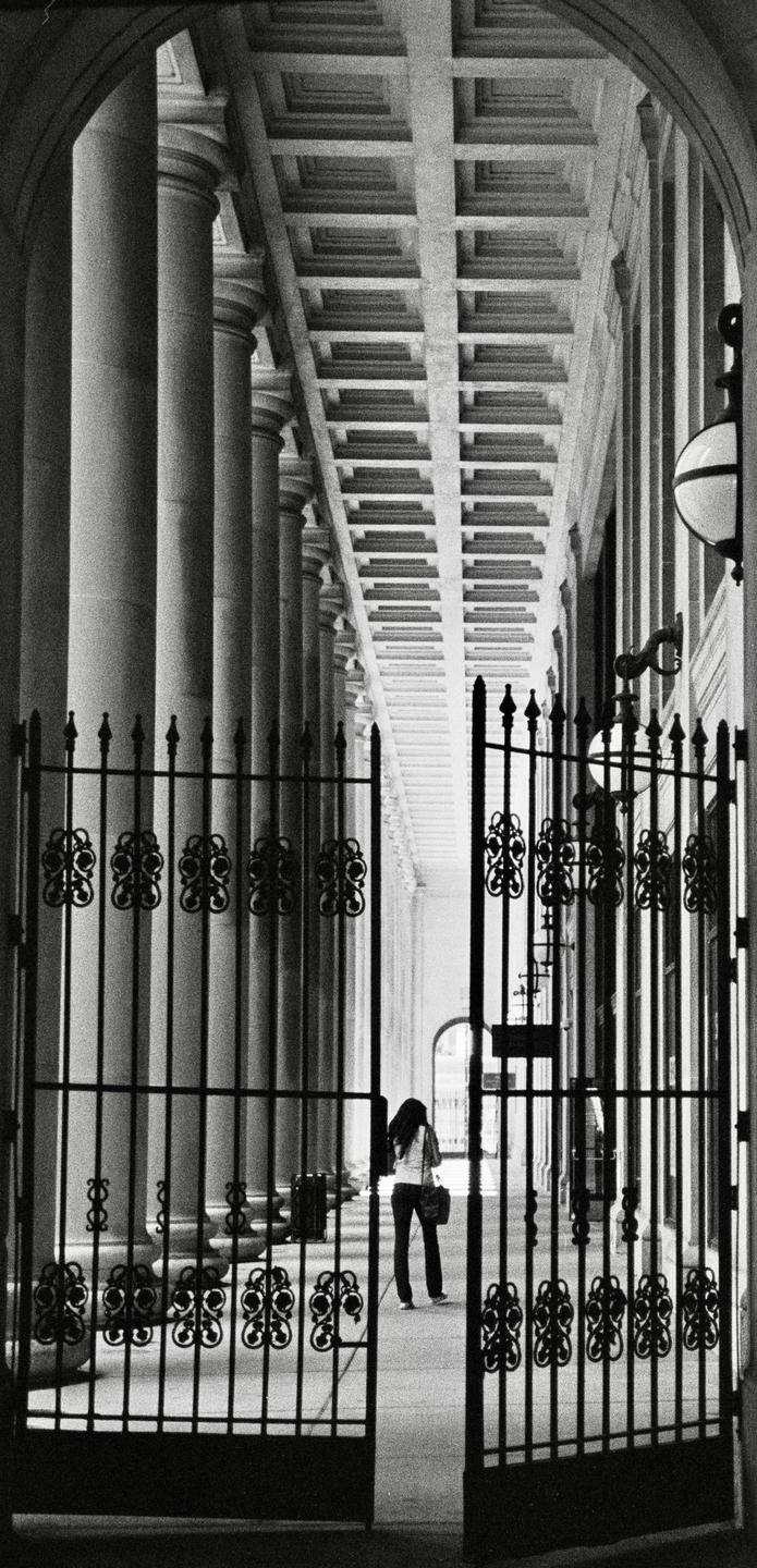 A woman walks away from the camera through an ornate gate into a long, columned hallway.