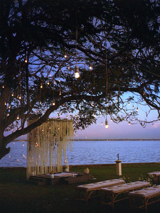 A wedding altar decorated with flowers and lights sits under a large tree by a lake at dusk.