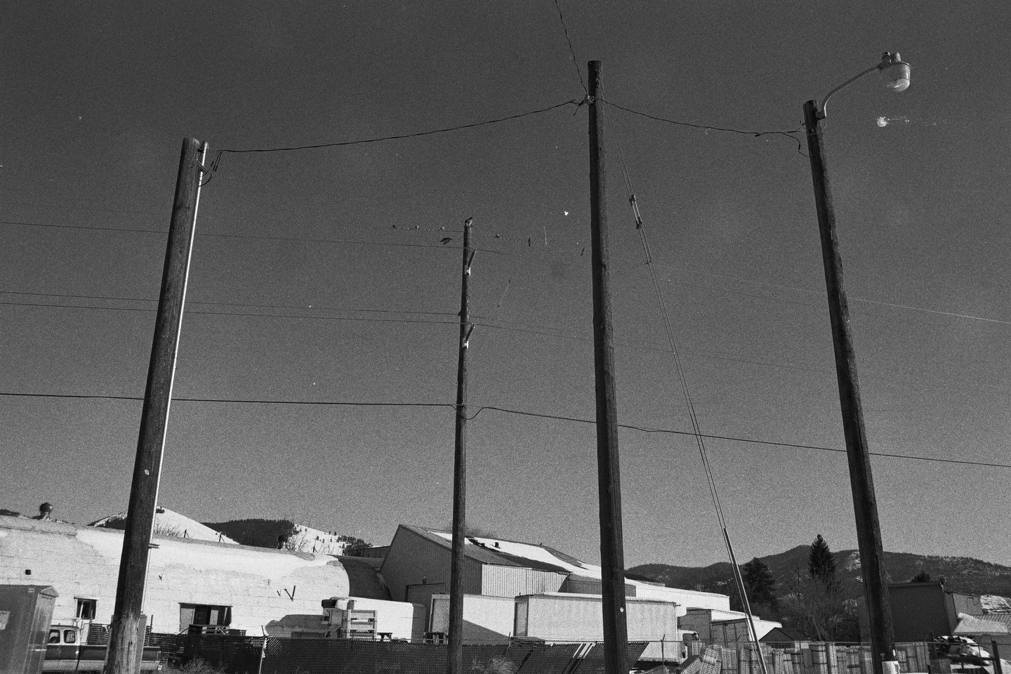 Four utility poles with power lines stand in front of industrial buildings and snow-covered hills under a clear sky.