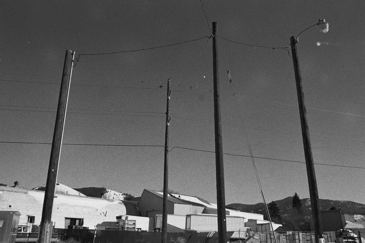 Four utility poles with power lines stand in front of industrial buildings and snow-covered hills under a clear sky.