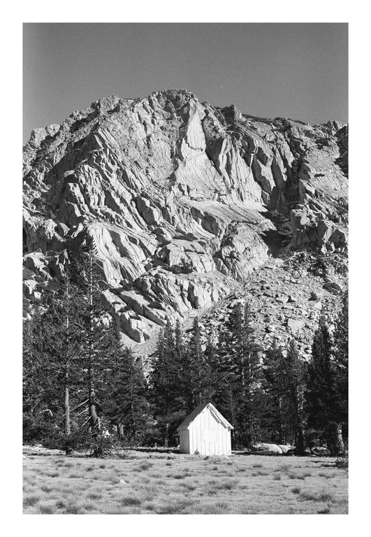 A small white cabin sits in a grassy field with a forest and a large mountain in the background.