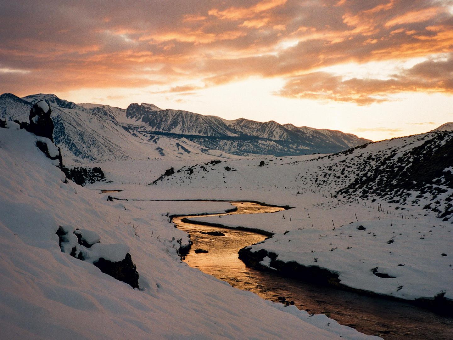 A winding river reflects the orange sky as it flows through a snow-covered mountain landscape.