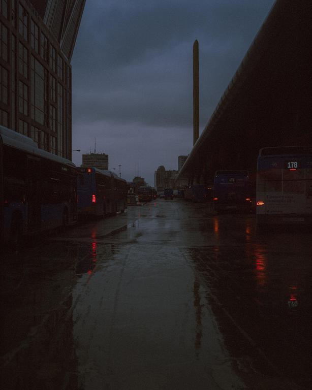 Buses are parked on a wet street next to a building under a dark, cloudy sky.