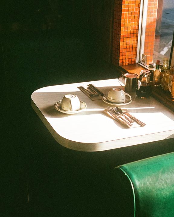 A diner table with cups, saucers, and cutlery is illuminated by a shaft of sunlight.