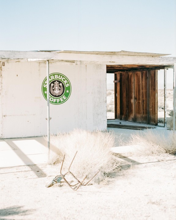 An abandoned building with a Starbucks logo on its white wall stands in a desert landscape with tumbleweeds and a metal frame on the ground.