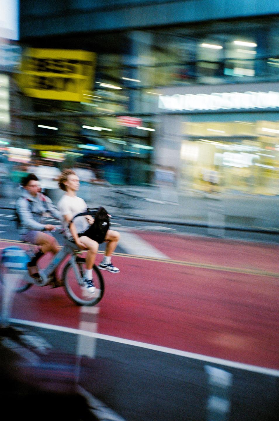 Two men ride a bicycle on a city street at dusk.