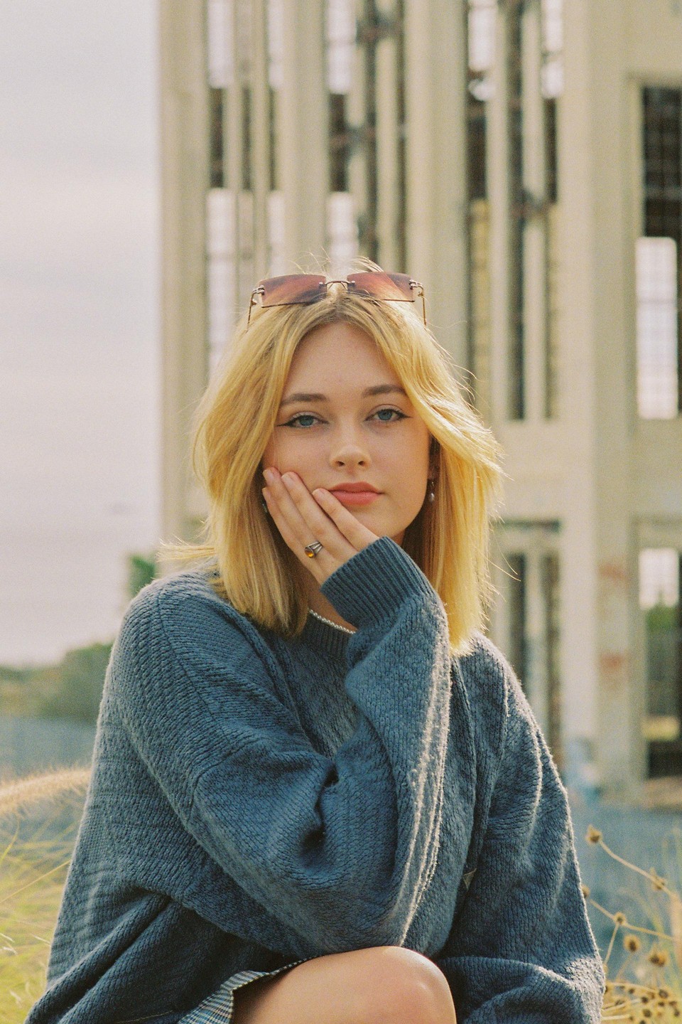 A woman with blonde hair and sunglasses on her head poses with her hand on her cheek in front of a building.